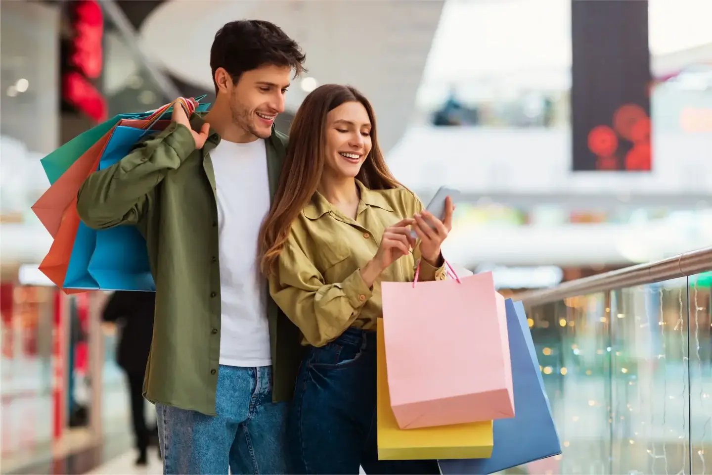 Smiling couple strolling with colorful shopping bags along a sunny open-air retail promenade lined with storefronts.