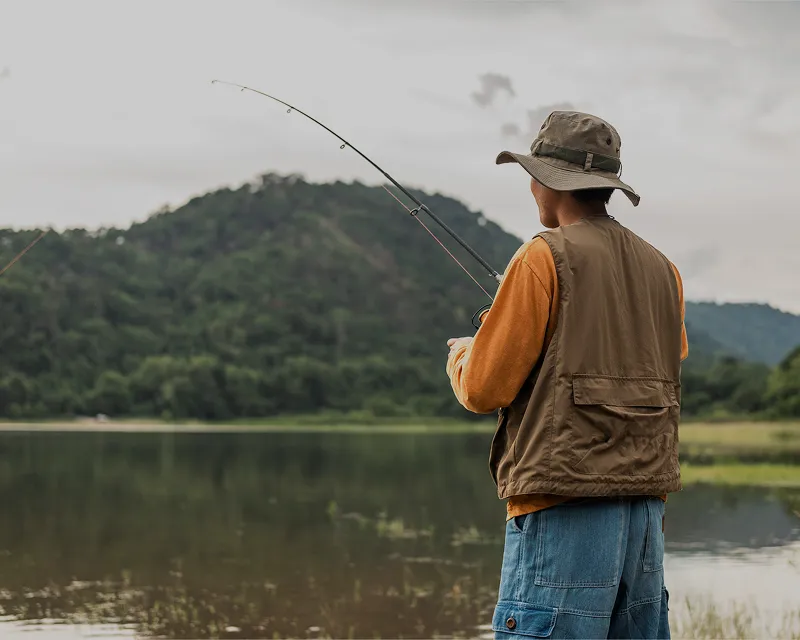 Man in a wide-brim hat casting a fishing line into a glassy lake with forested mountains rising in the far distance.