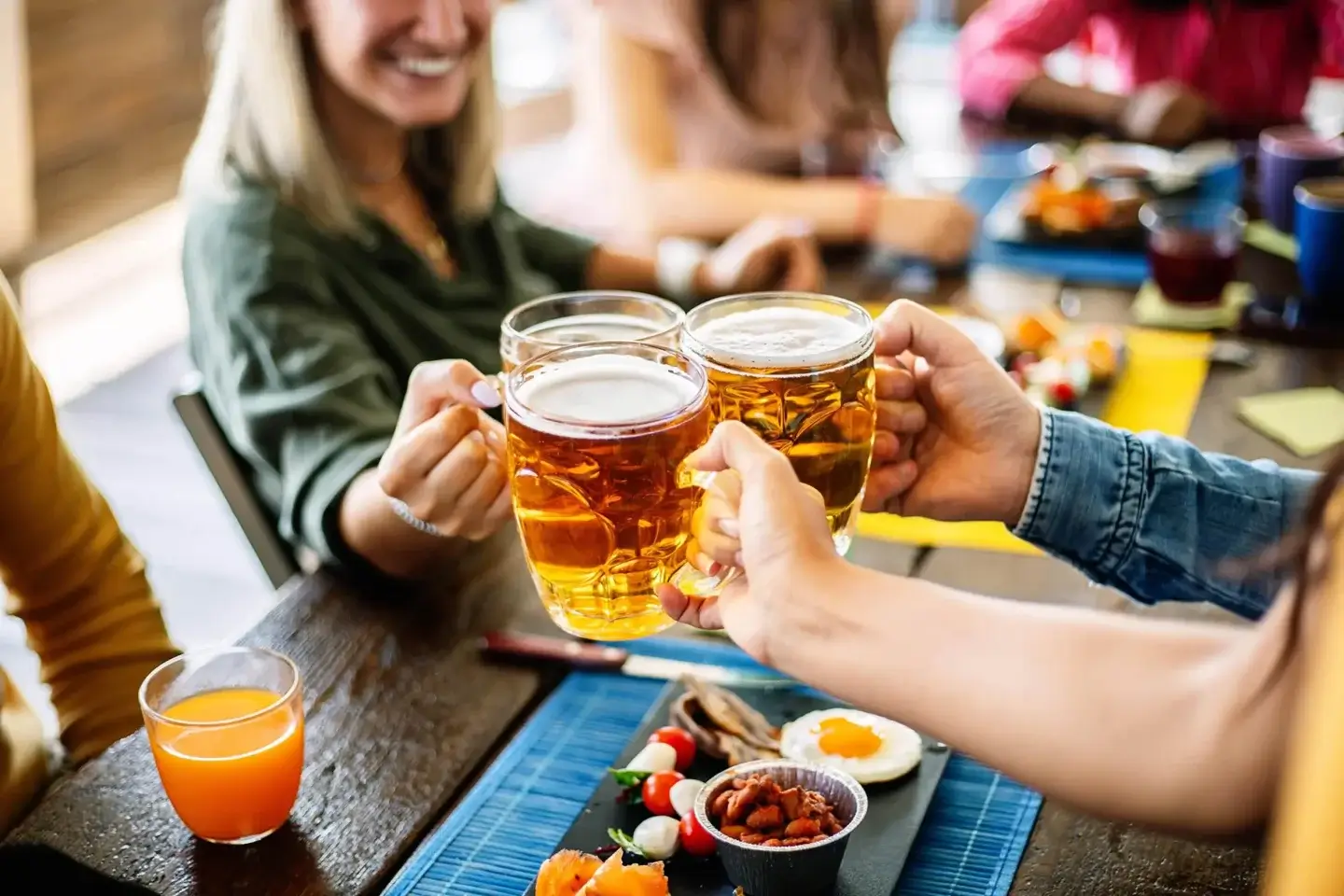 Three people clinking tall glasses of golden beer above a wooden table set with a shared plate of appetizers.