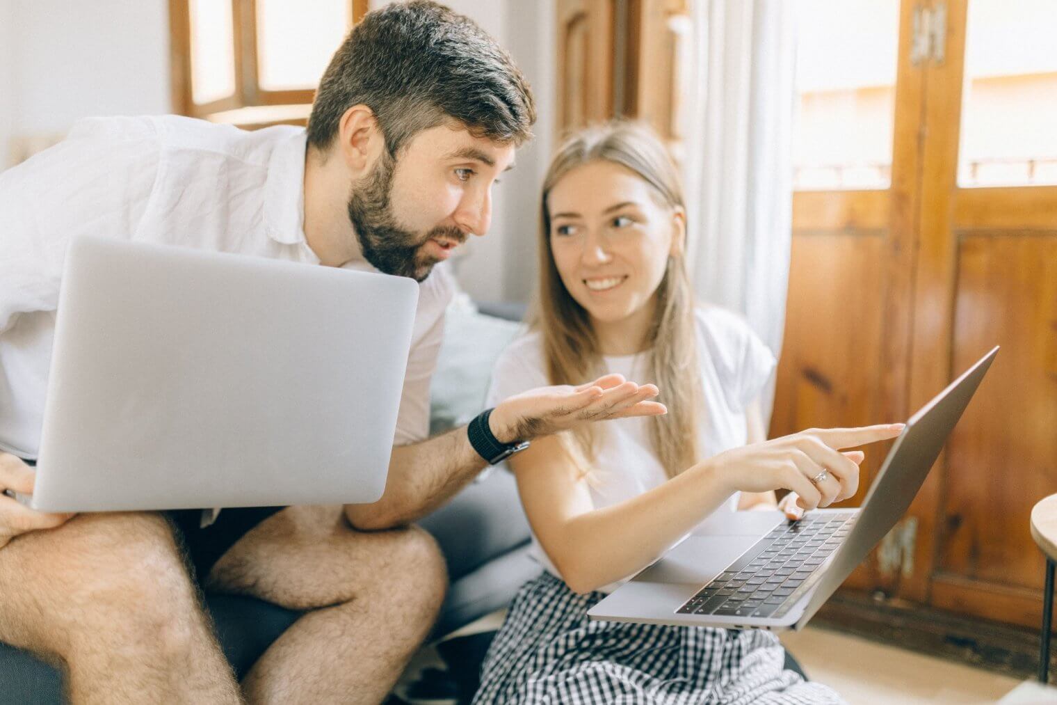A man holding a laptop leaning toward a woman sitting on the floor pointing at her laptop screen inside a room. 