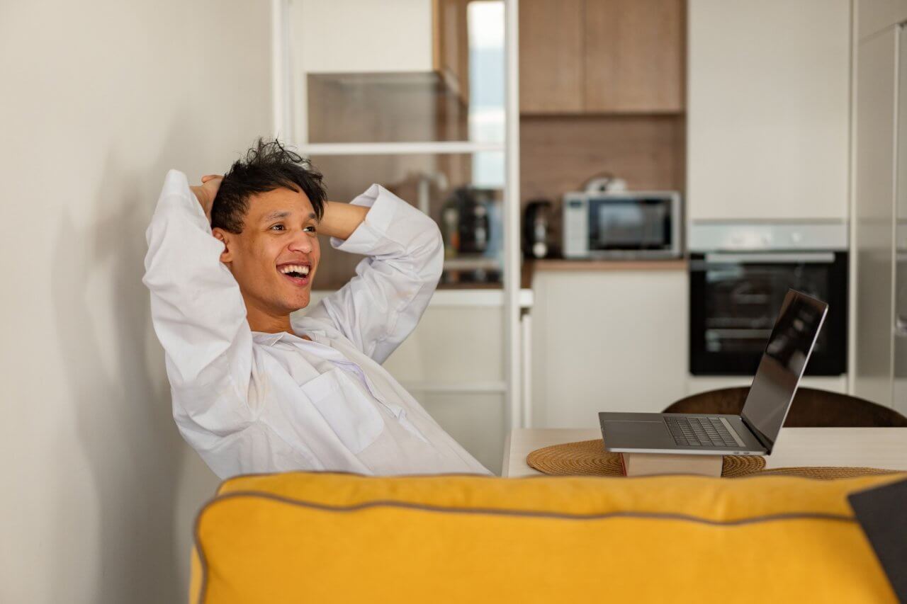 2 pexels-mikhail-nilov-7593804 A smiling man sitting at a table leaning back with hands behind his head looking at a laptop in front of a kitchen.