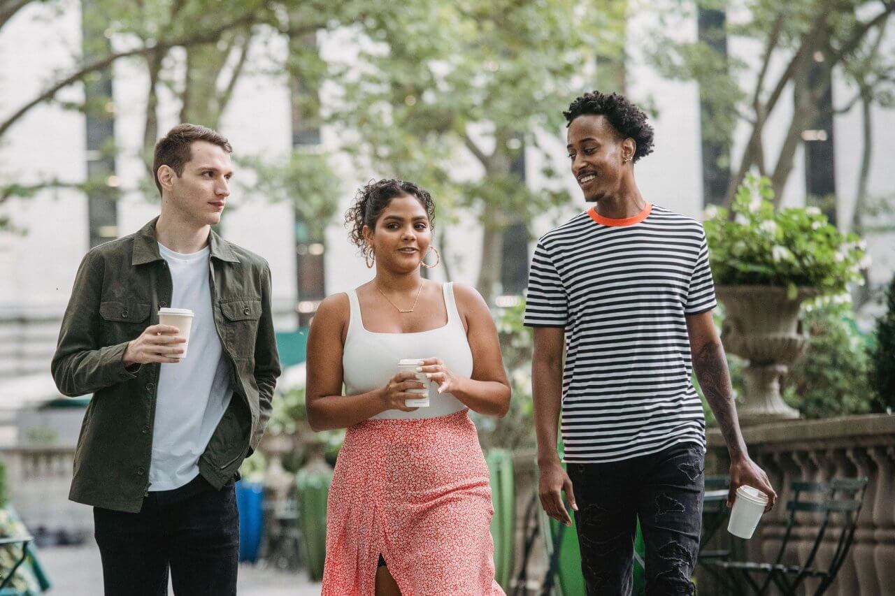 2 pexels-william-fortunato-6140382 Three diverse young friends holding takeaway coffee cups while walking and chatting together outdoors in a city park.