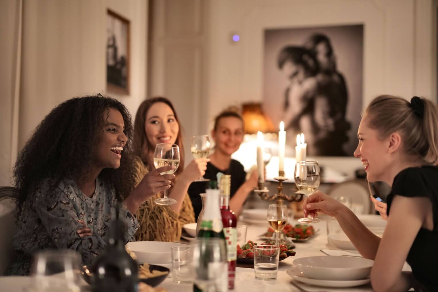 Four friends laughing and toasting with wine glasses around a candlelit dinner party table in an apartment.